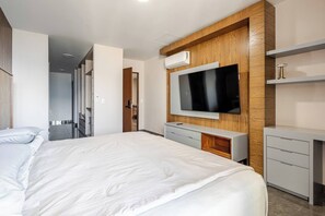A modern hotel room with a large bed, a TV mounted on a wooden panel, a small desk, and a wardrobe.