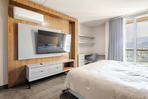 A modern hotel room with a large bed, a mounted TV, a desk, and a view of the city through large windows.