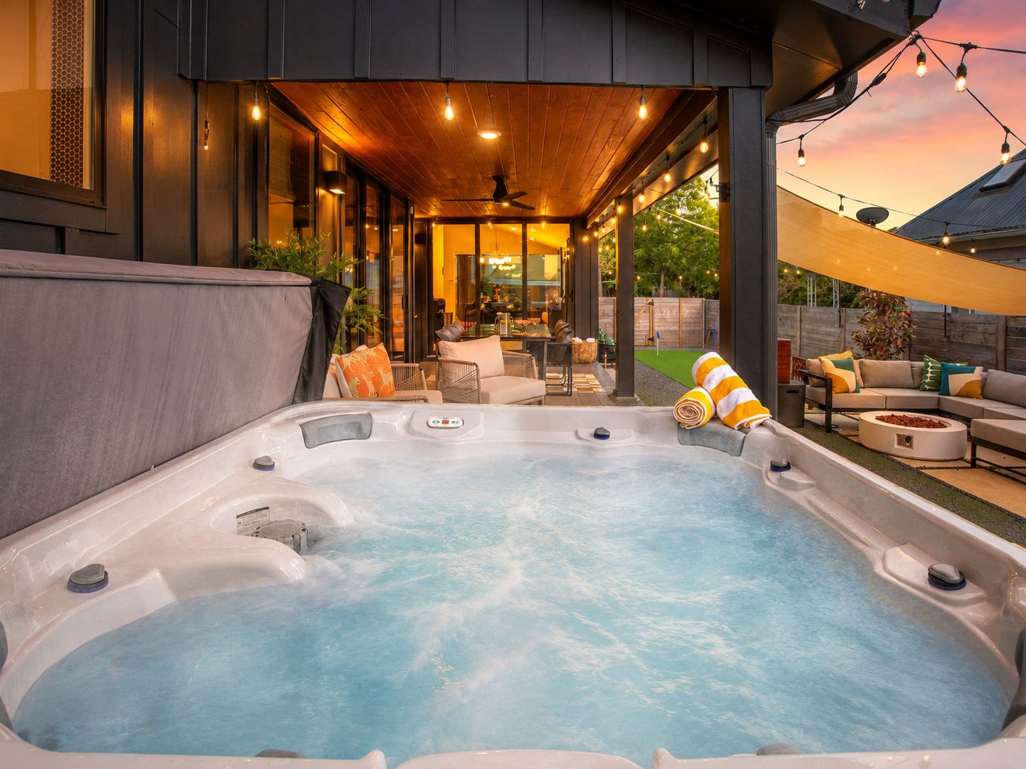 A hot tub with a wooden deck and outdoor furniture, surrounded by a covered area with string lights.