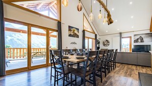Uma sala de jantar espaçosa com soalho em madeira, uma mesa comprida e cadeiras modernas. Grandes janelas oferecem vista para o exterior.