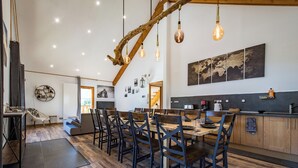 Uma ampla área de refeições com uma longa mesa de madeira e cadeiras, teto rústico com vigas aparentes e uma cozinha moderna com bancada preta e armários de madeira.