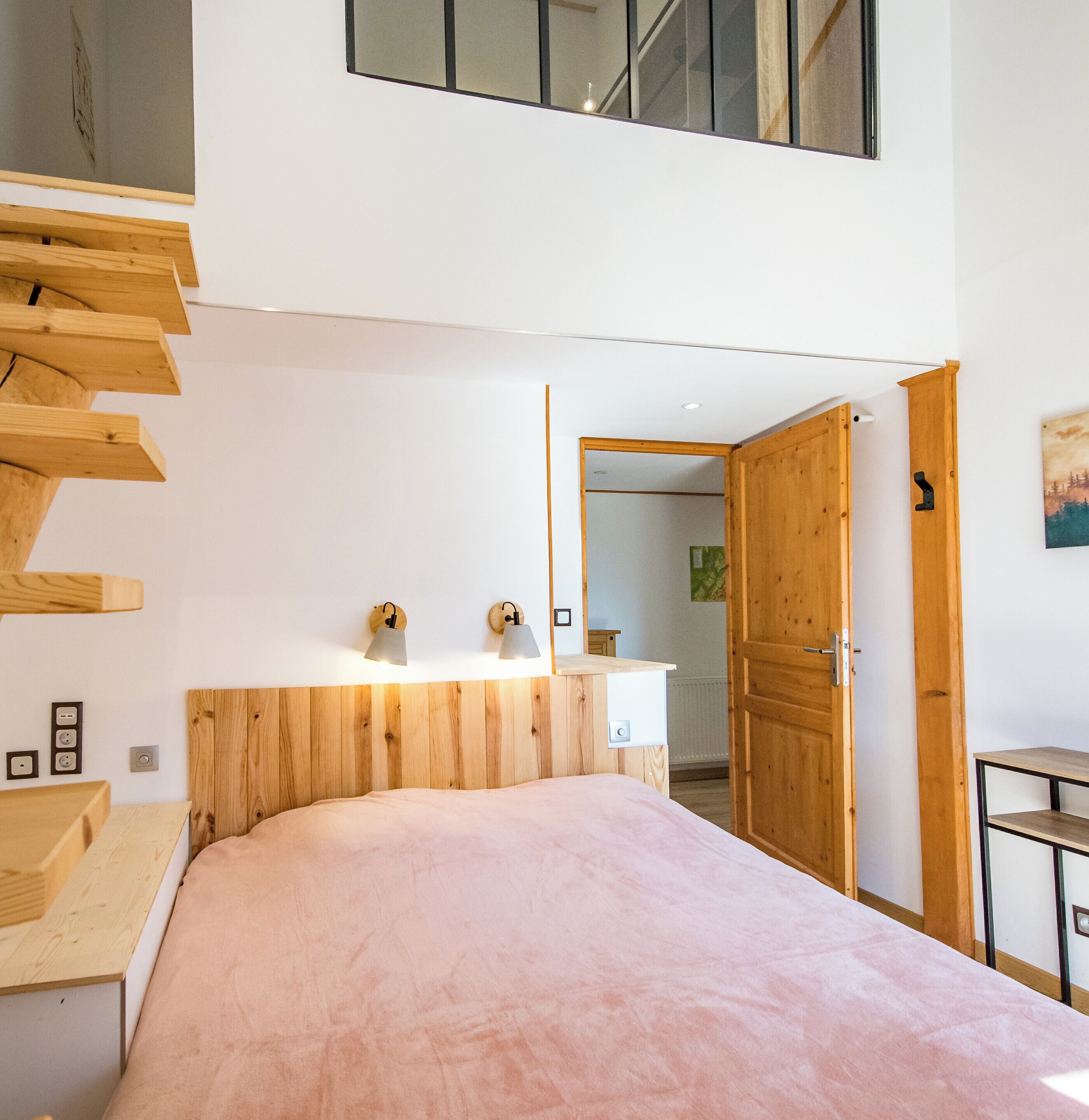 Ein Schlafzimmer mit hölzernem Kopfteil, einem Bett mit rosa Bettdecke, einem Schreibtisch mit Stuhl, einem Holzschrank und einem kleinen Tisch.