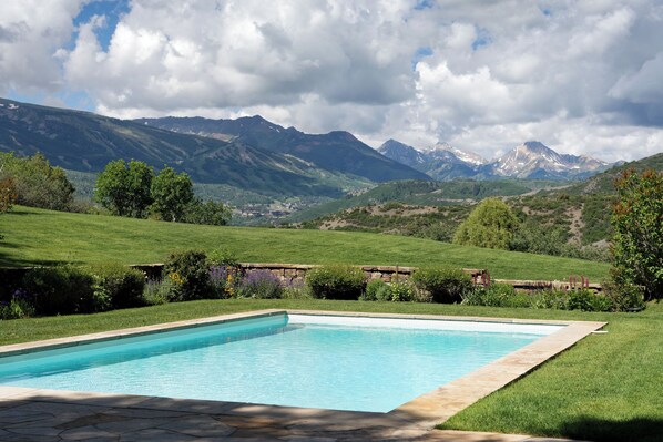 Une piscine rectangulaire bordée de pierres, entourée d’une pelouse bien entretenue et d’un mur de pierres, le tout se dressant sur un fond de montagnes et d’un ciel nuageux.