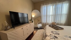 Een slaapkamer met een flatscreen televisie op een kast, een bed met bloemenbedrukking, een nachtkastje met een dienblad en een raam met dunne gordijnen.