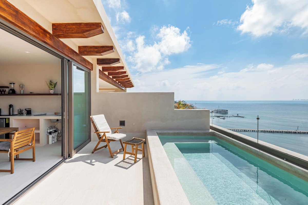 A modern balcony with a pool, wooden furniture, and a view of the sea.