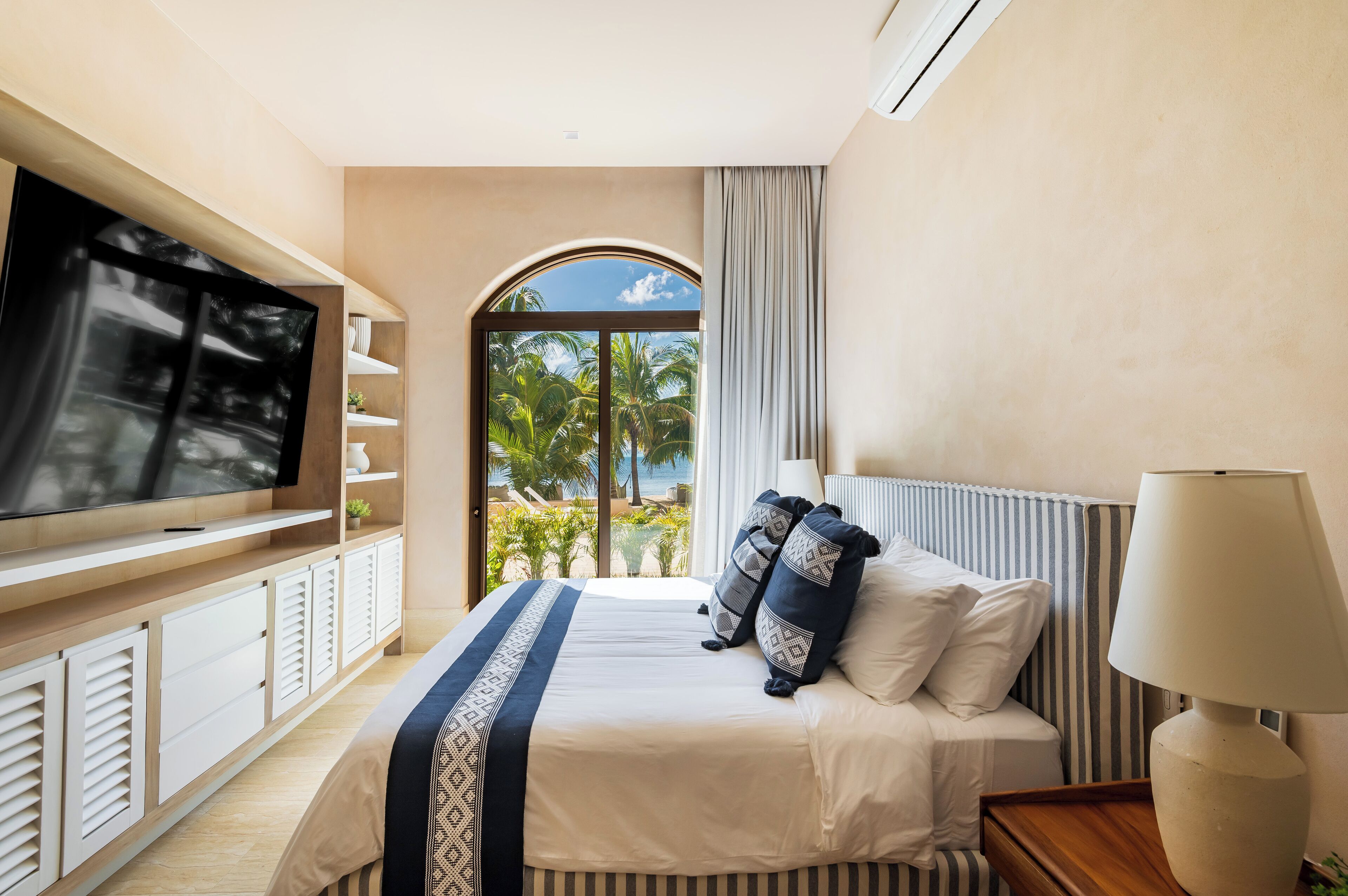 A hotel room with a large flat-screen TV, a bed with white and patterned pillows, a bedside lamp, and a view of palm trees through a window.