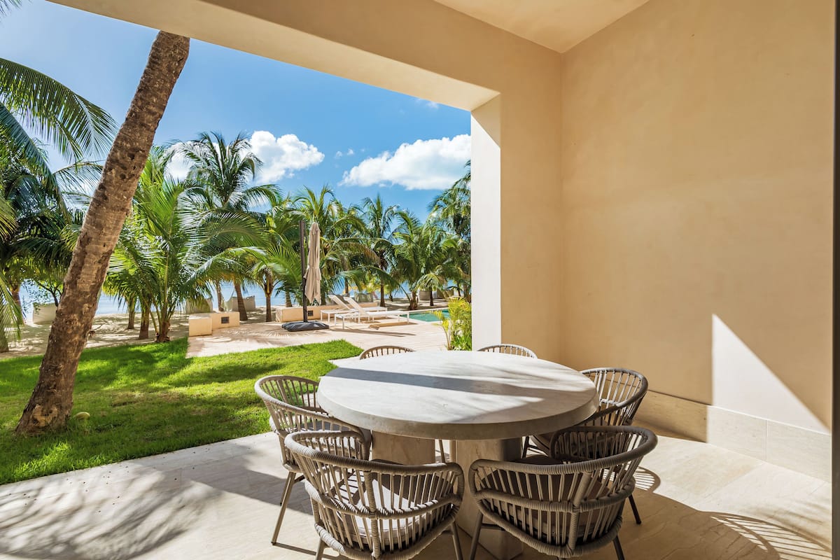 An outdoor patio with a round table and wicker chairs, overlooking a pool area with palm trees and lounge chairs.