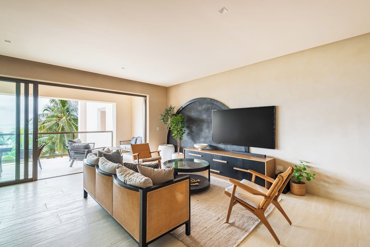 A modern living room with a sofa, chairs, a coffee table, a TV, and a balcony with a view of palm trees.