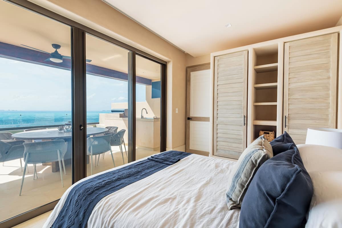 A bedroom with a bed, a balcony with a table and chairs, and a view of the sea.