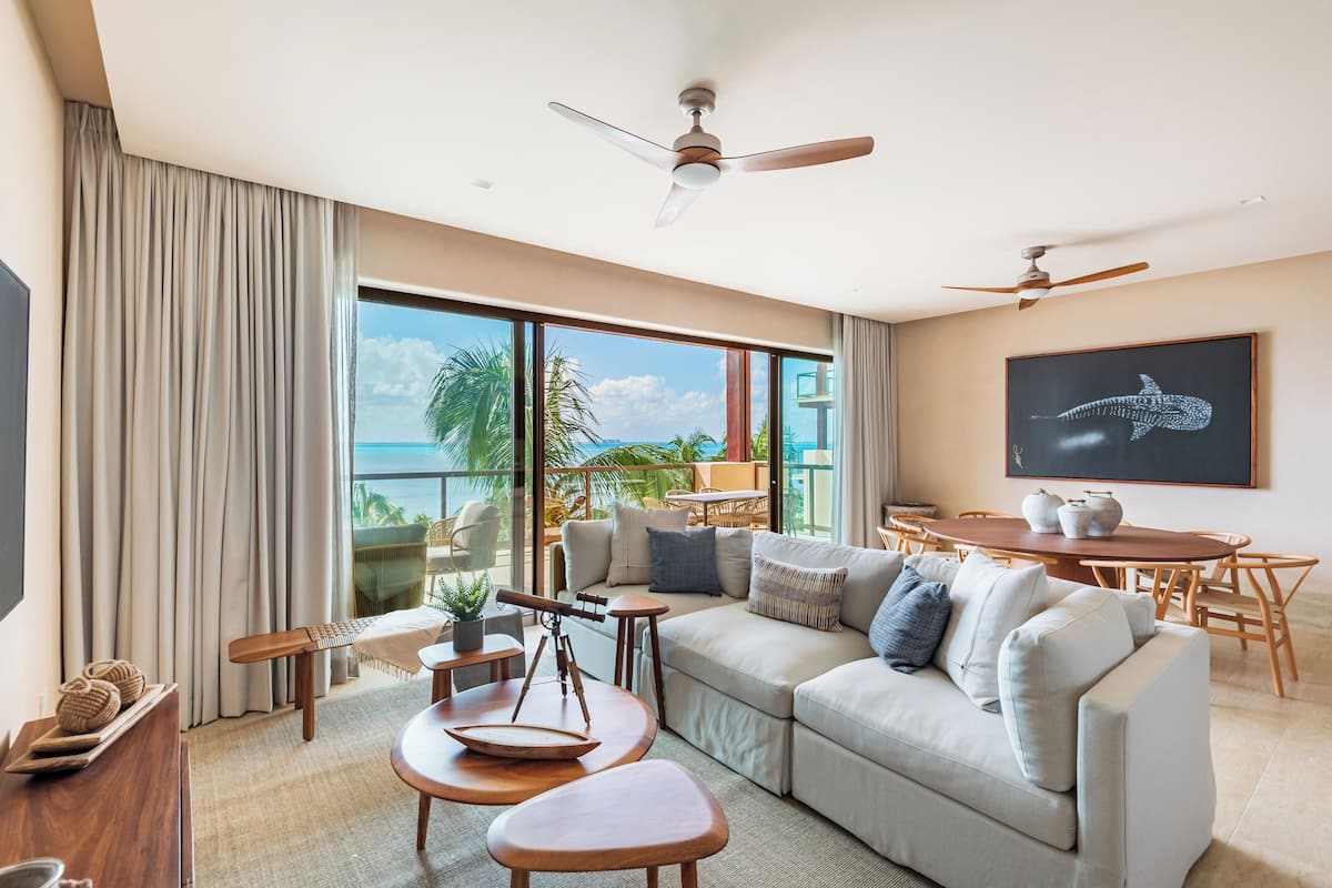 A modern living room with a large sofa, wooden coffee table, and a balcony with a view of palm trees and the ocean.