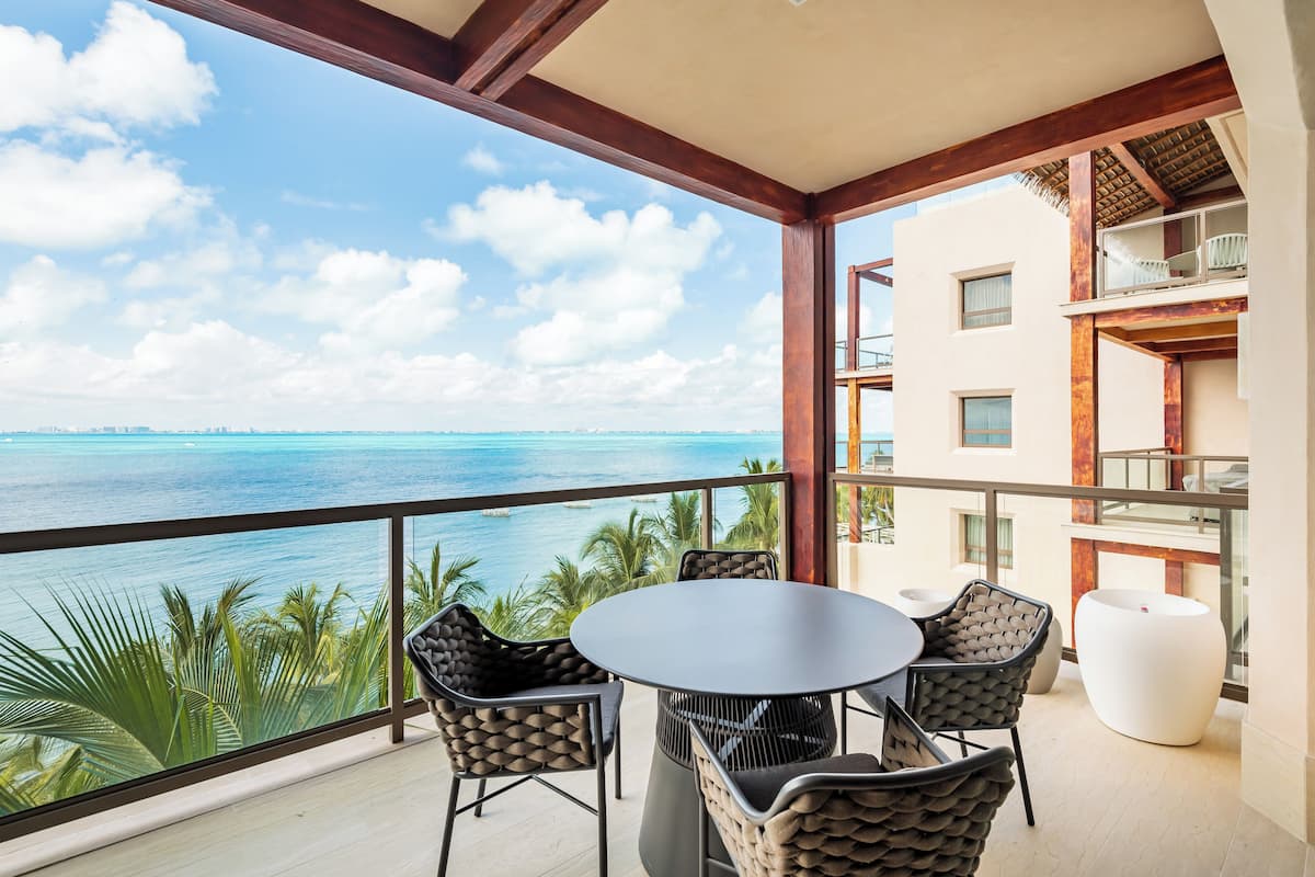 A balcony with a round table and chairs, overlooking a turquoise sea and palm trees.