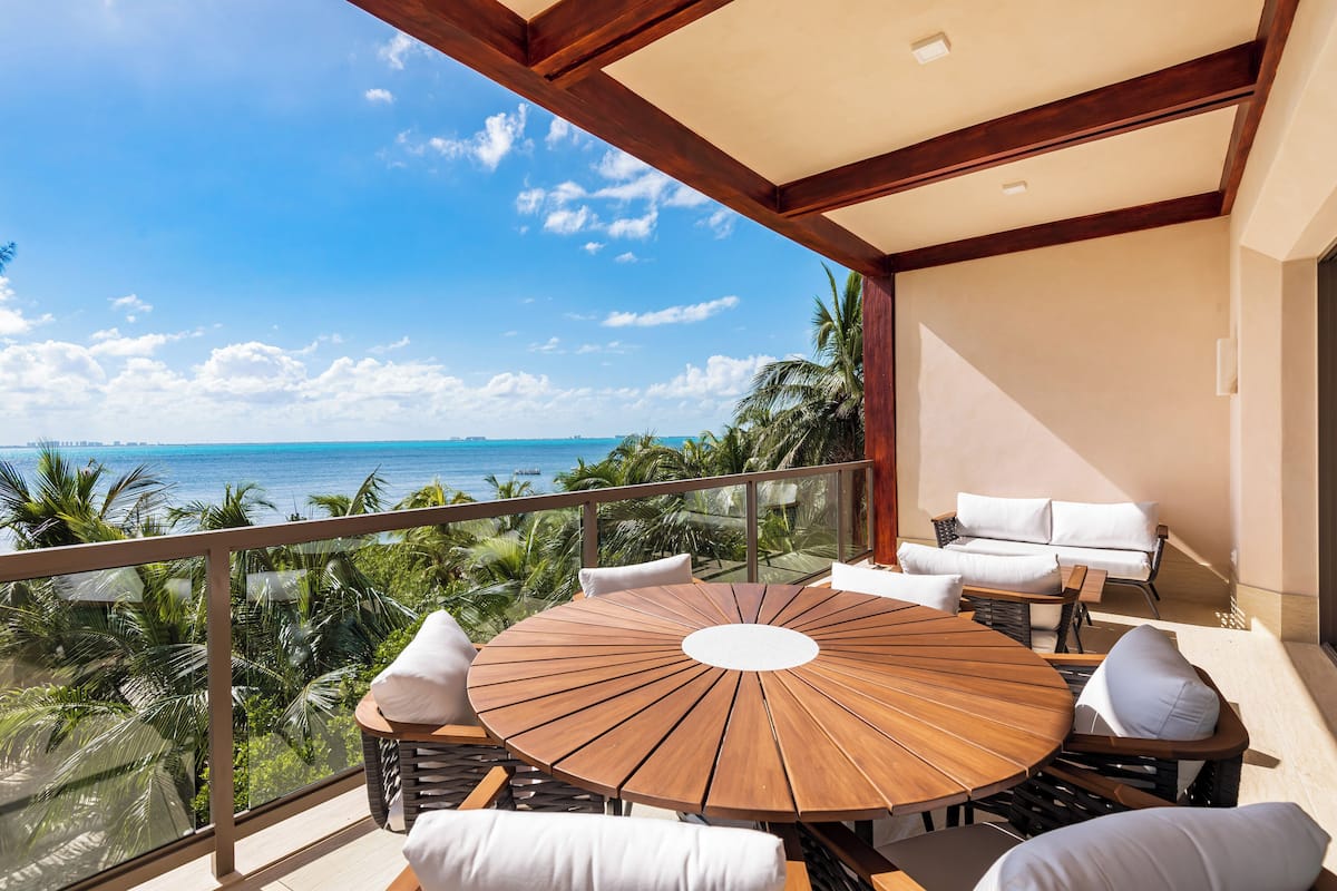 A round wooden table with white cushions on a balcony overlooking a beach with palm trees and the ocean.