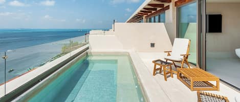 Un área de piscina moderna al aire libre con vista despejada al océano, sillones de madera para tomar el sol y una sala con paredes de vidrio.