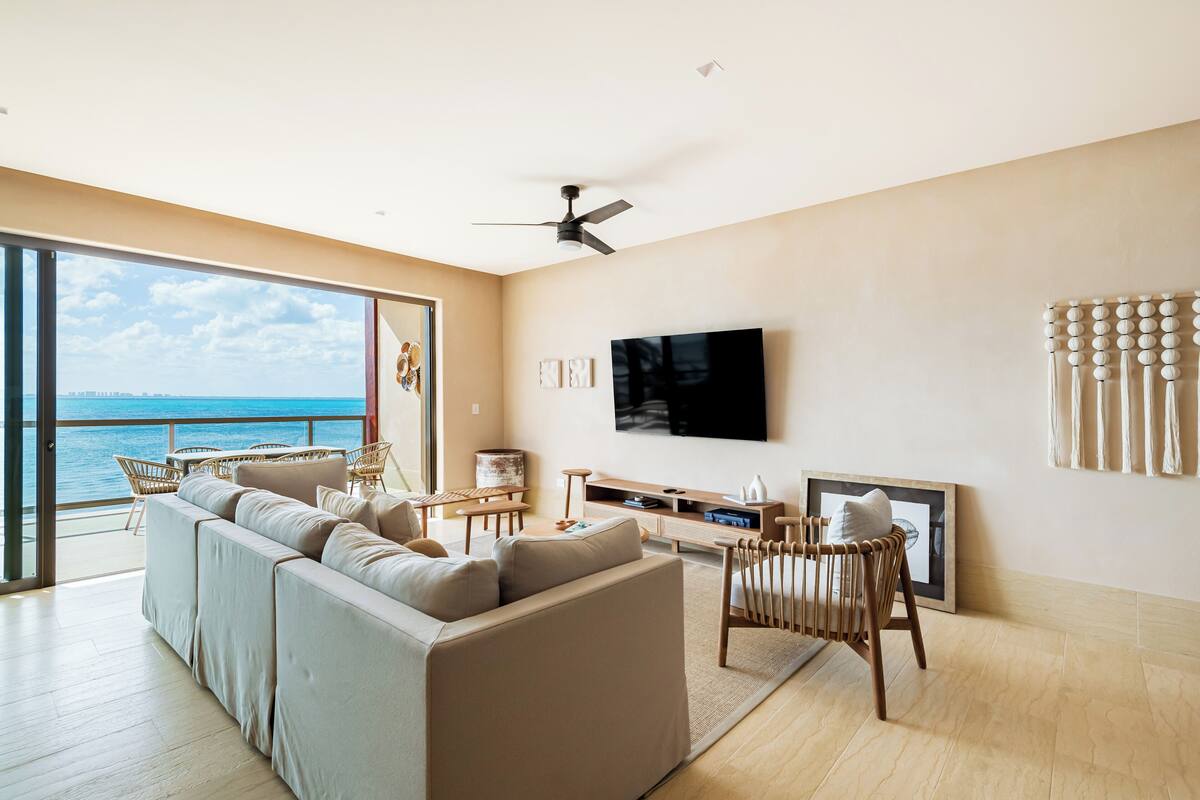 A modern living room with a large sofa, a flat-screen TV, and a view of the ocean.