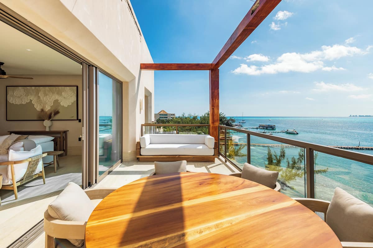 A modern outdoor balcony with a wooden table and chairs, a white sofa, and a view of the ocean and boats.