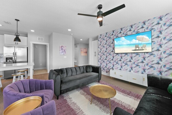 Un salon moderne avec un canapé gris, un fauteuil violet et une table basse. On y trouve un ventilateur de plafond, un coin cuisine avec des meubles blancs et un grand tableau représentant une scène de plage accroché au mur.
