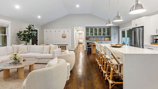 A spacious living room with a white sofa, a coffee table, and a large plant. A kitchen with white cabinets and stainless steel appliances is visible in the background. - Upscale Secluded Home w/Central AC, Pool & Firepit (Centerville,)