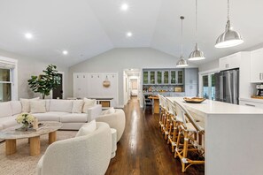 A spacious living room with a white sofa, a coffee table, and a large plant. A kitchen with white cabinets and stainless steel appliances is visible in the background.