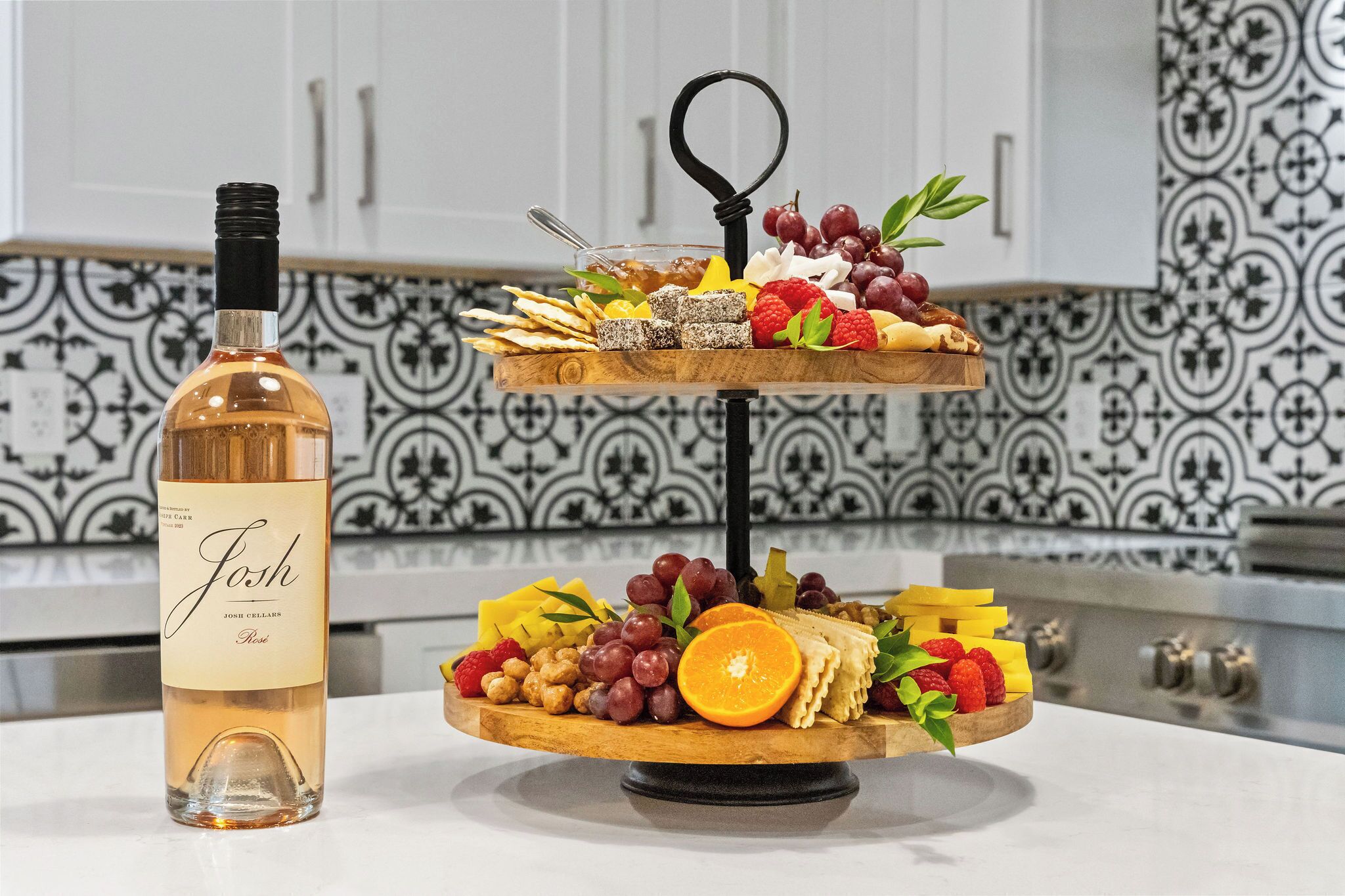 A two-tiered wooden board with various cheeses, fruits, and crackers, accompanied by a bottle of wine.