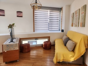 A room with a wooden floor, a yellow sofa, a small table with a plant, and a window with blinds.