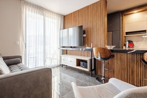 Un salon moderne comprenant un canapé, un mur d’accent en bois, un téléviseur et un coin cuisine avec un comptoir et des tabourets de bar.