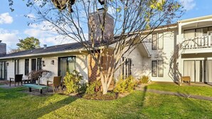 Uma casa de dois andares com chaminé de tijolos, varanda frontal com banco e um pequeno jardim.