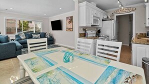 A modern living room with a blue sofa, a dining table with a beach-themed table runner, and a kitchen area with white cabinets and a welcome sign.