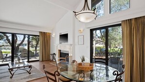 A spacious living room with a glass table, comfortable seating, a fireplace, and large windows offering a view of the outdoors.