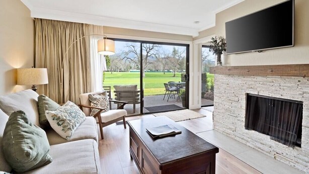 Un salon moderne avec un canapé, une table basse et une cheminée. Un téléviseur est fixé au mur, et une grande fenêtre offre une vue sur un paysage verdoyant.