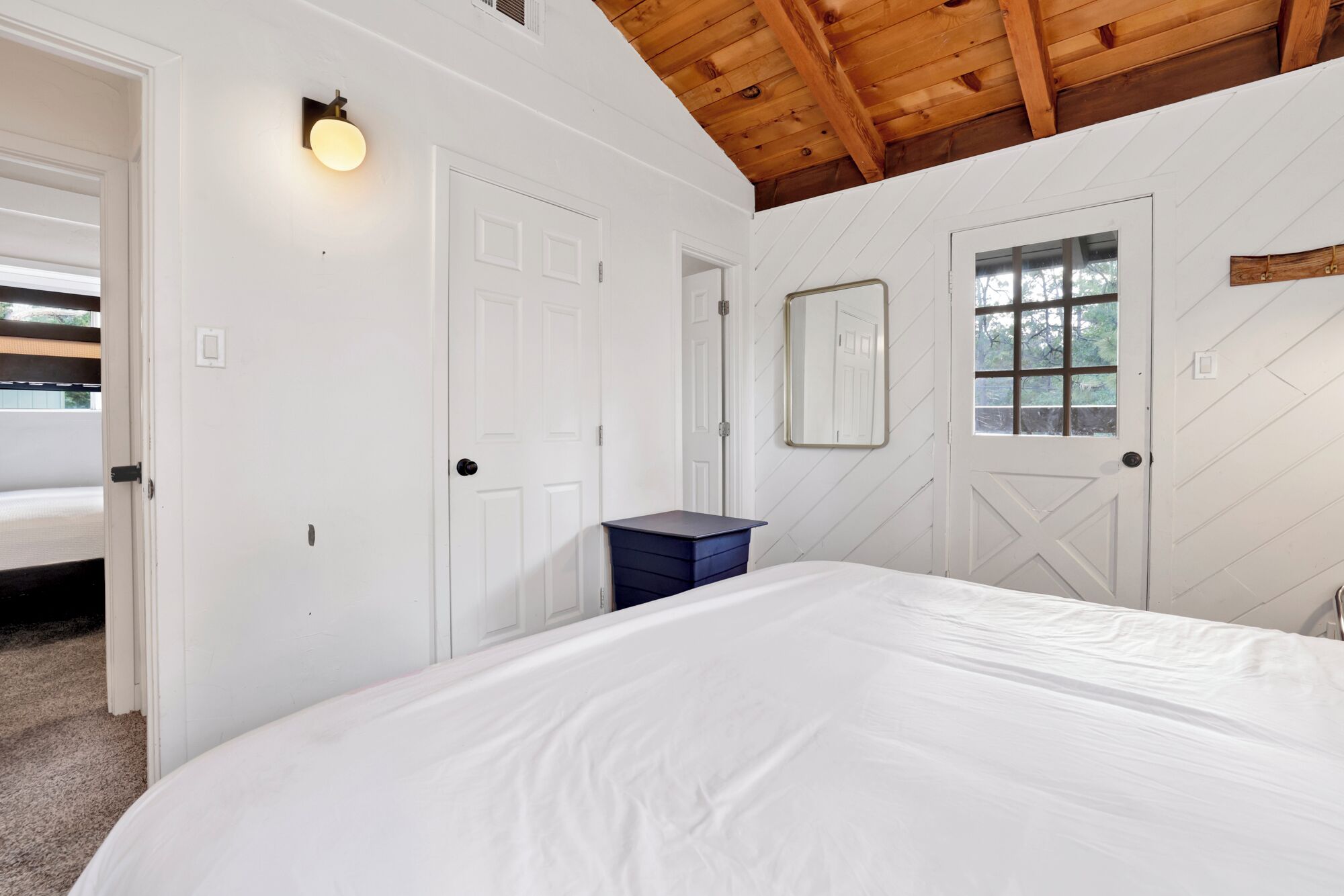 A bedroom with a large bed, white walls, a wooden ceiling, and a window with a view of trees.