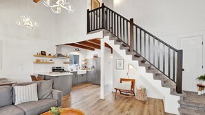 A spacious living area with a grey sofa, wooden coffee table, and a book. A staircase with a black railing leads to a kitchen area with a fridge and a dining table. - Large Bear Lodge #2488 (Sugarloaf)