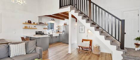 Amplia sala de estar con un sofá gris, una mesa de centro de madera y un libro. Una escalera con barandilla negra lleva a una zona de cocina con un refrigerador y una mesa de comedor.