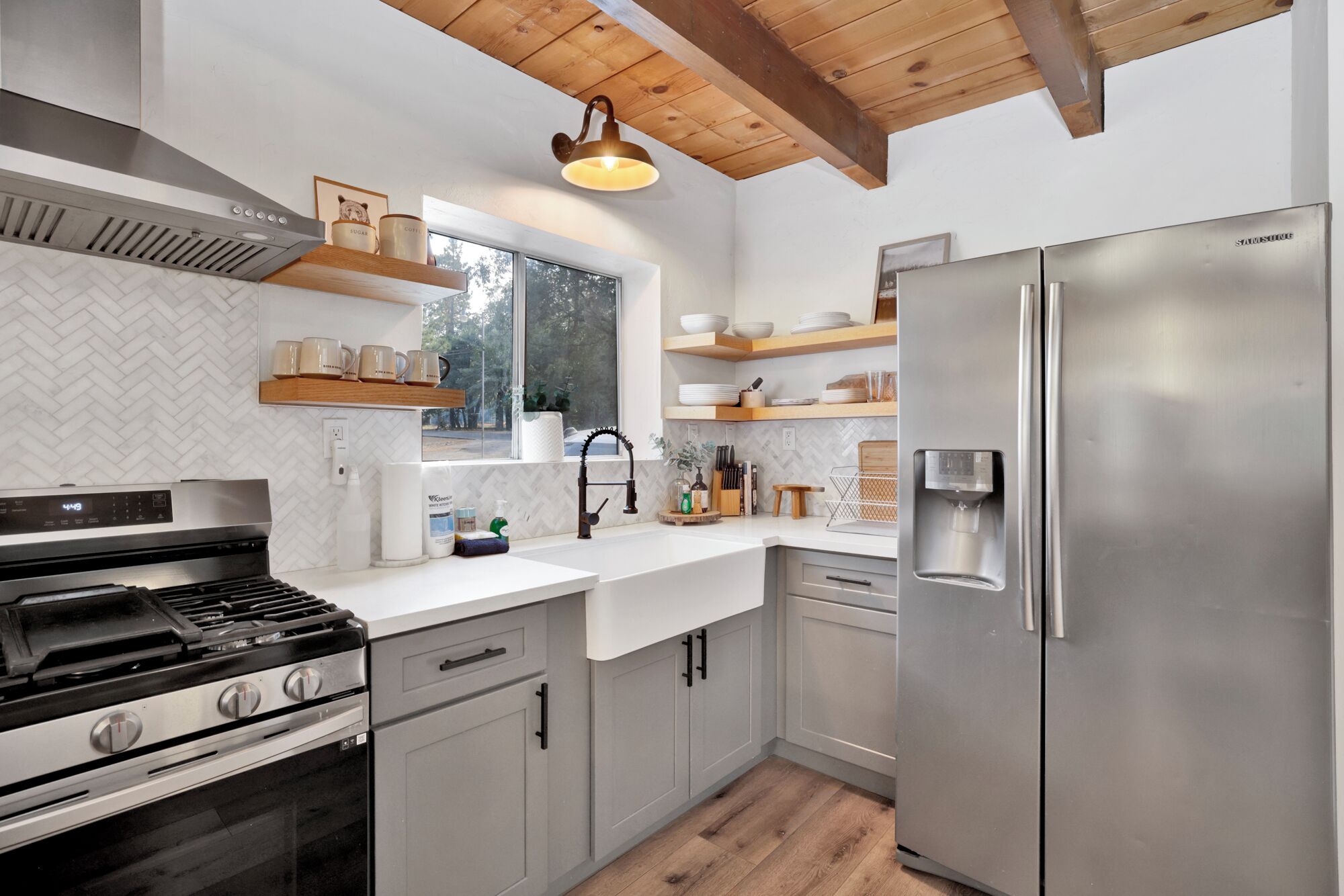 Une cuisine moderne dotée d’un réfrigérateur en acier inoxydable, d’une cuisinière à gaz, d’étagères en bois et d’une fenêtre donnant sur les arbres.