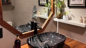 Een badkamer met een houten wastafelmeubel, een zwarte marmeren wastafel, een grote spiegel en kunstwerken in lijsten aan de muur.