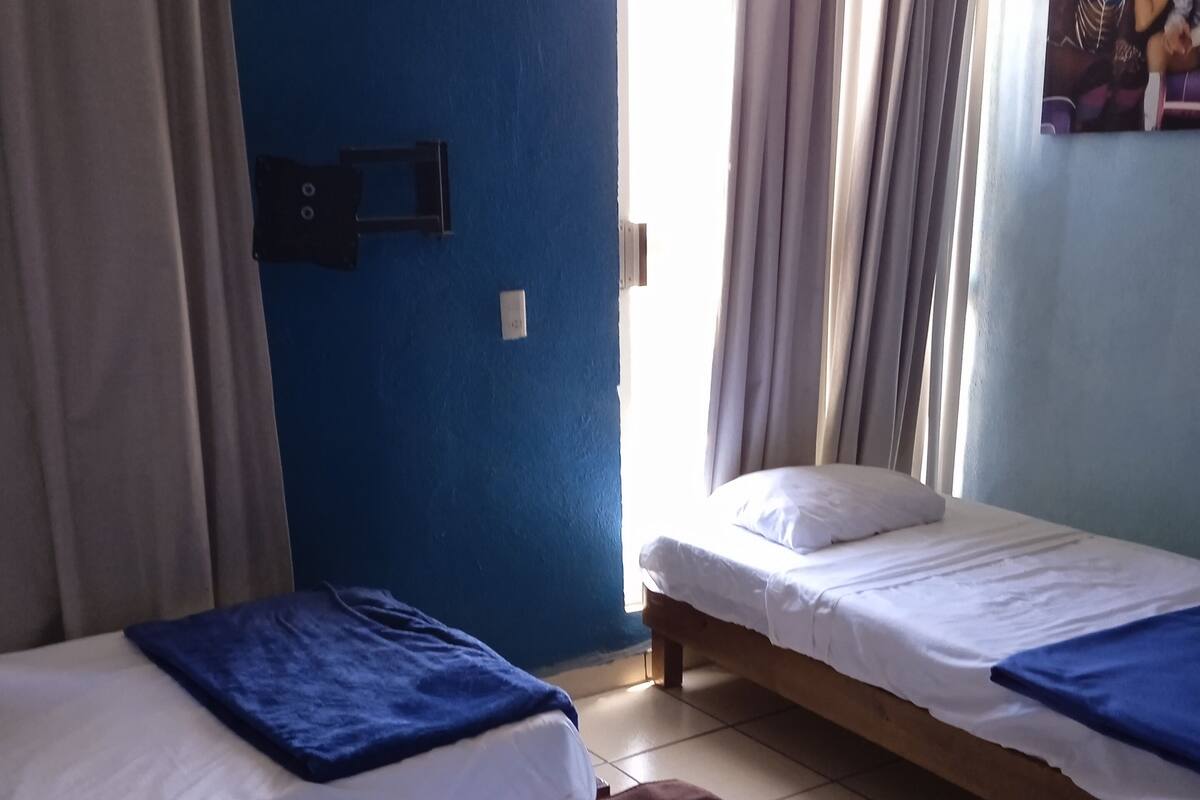 A room with two beds, each with a blue and white blanket. There is a window with curtains, and a wall-mounted light switch.