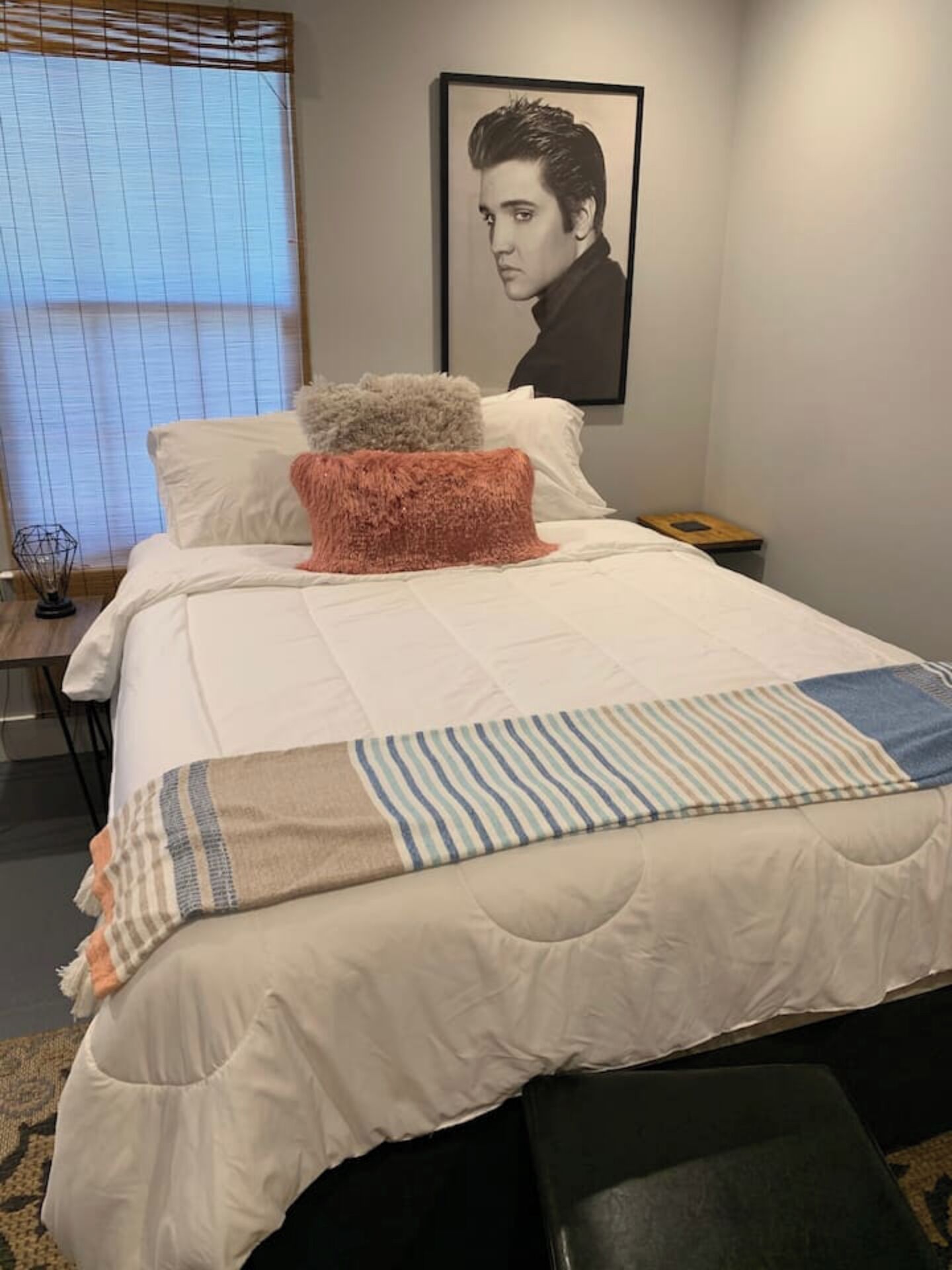 Ein ordentlich bezogenes Bett mit einer gestreiften Decke und einem flauschigen Kopfkissen, ein gerahmtes Bild an der Wand und ein kleiner Nachttisch.