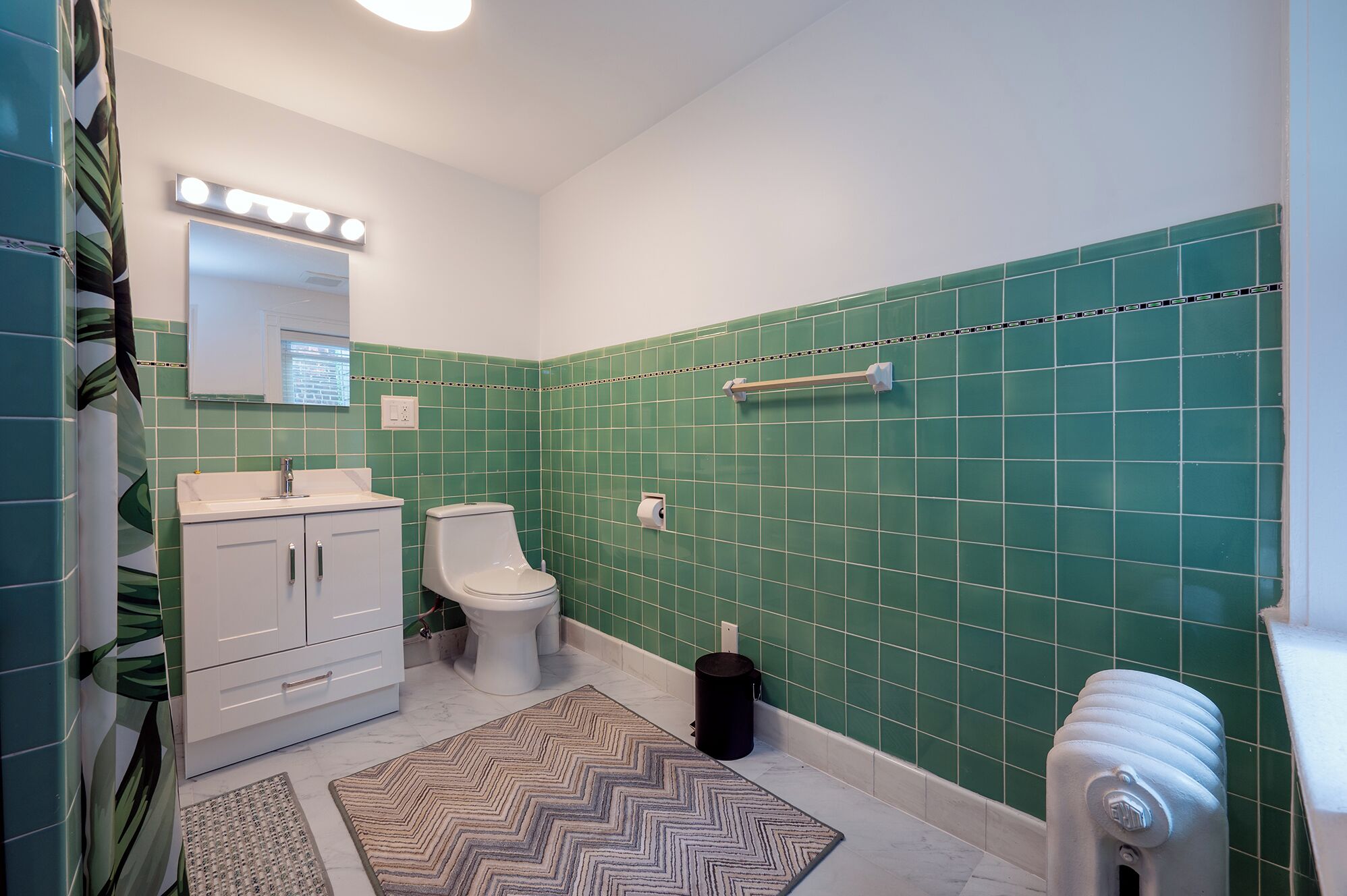 A bathroom with a white sink, toilet, and cabinet, green tiled walls, and a mirror with lights above.