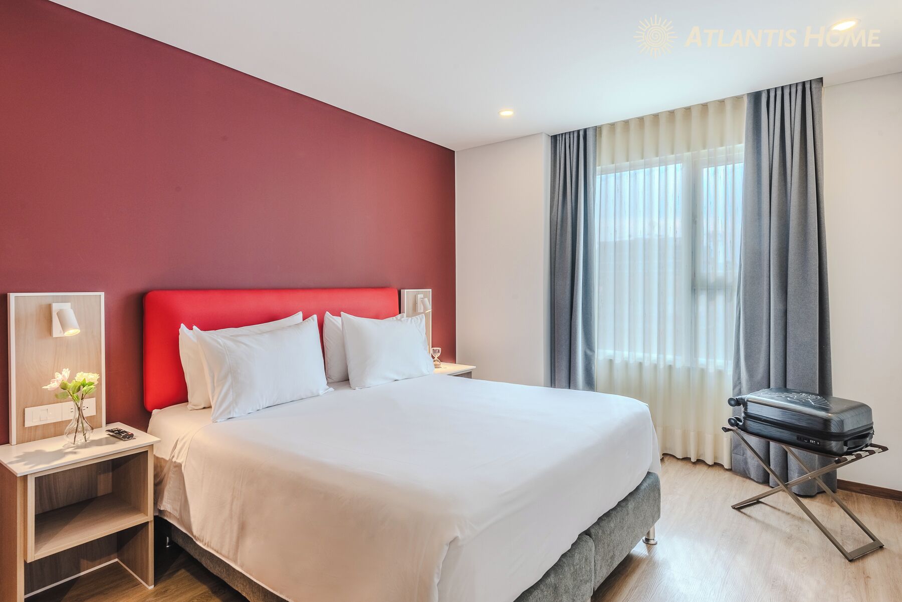 Habitación de hotel con una cama grande, una mesita de noche con un jarrón y control remoto, una maleta y una ventana con cortinas.