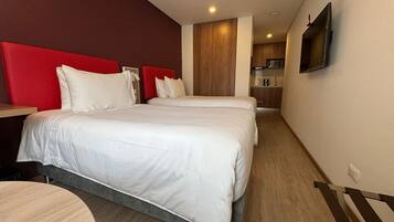 Una habitación de hotel con dos camas, un televisor montado en la pared y una kitchenette al fondo.