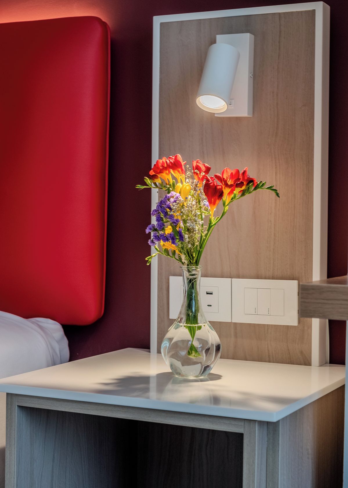 Habitació d'hotel moderna amb una paret vermella decorativa, una taula de nit blanca i un vas transparent amb flors de colors.