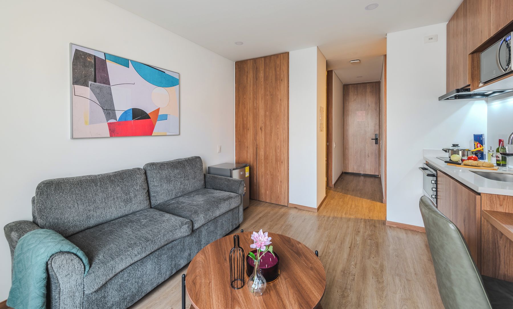 Un salon moderne avec un canapé gris, une table à manger en bois agrémentée d’un vase et d’une bouteille, et un coin cuisine doté de meubles et d’un plan de travail en bois.