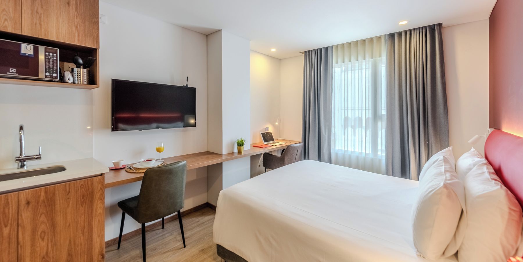 A hotel room with a bed, a desk with a computer, a TV mounted on the wall, and a window with curtains.
