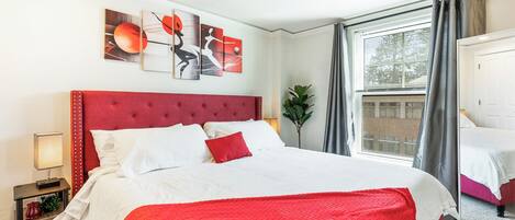 Une chambre d’hôtel avec une tête de lit rembourrée en tissu rouge, un grand lit avec une literie blanche et rouge, une table de chevet avec une lampe et une fenêtre avec des rideaux.