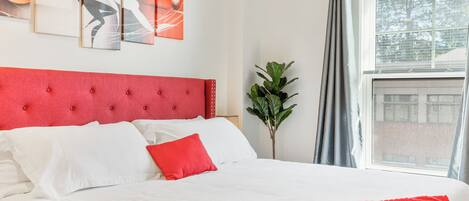 Een bed met een rode hoofdbord, witte beddengoed en een rode plaid. Boven het bed hangen drie abstracte schilderijen.