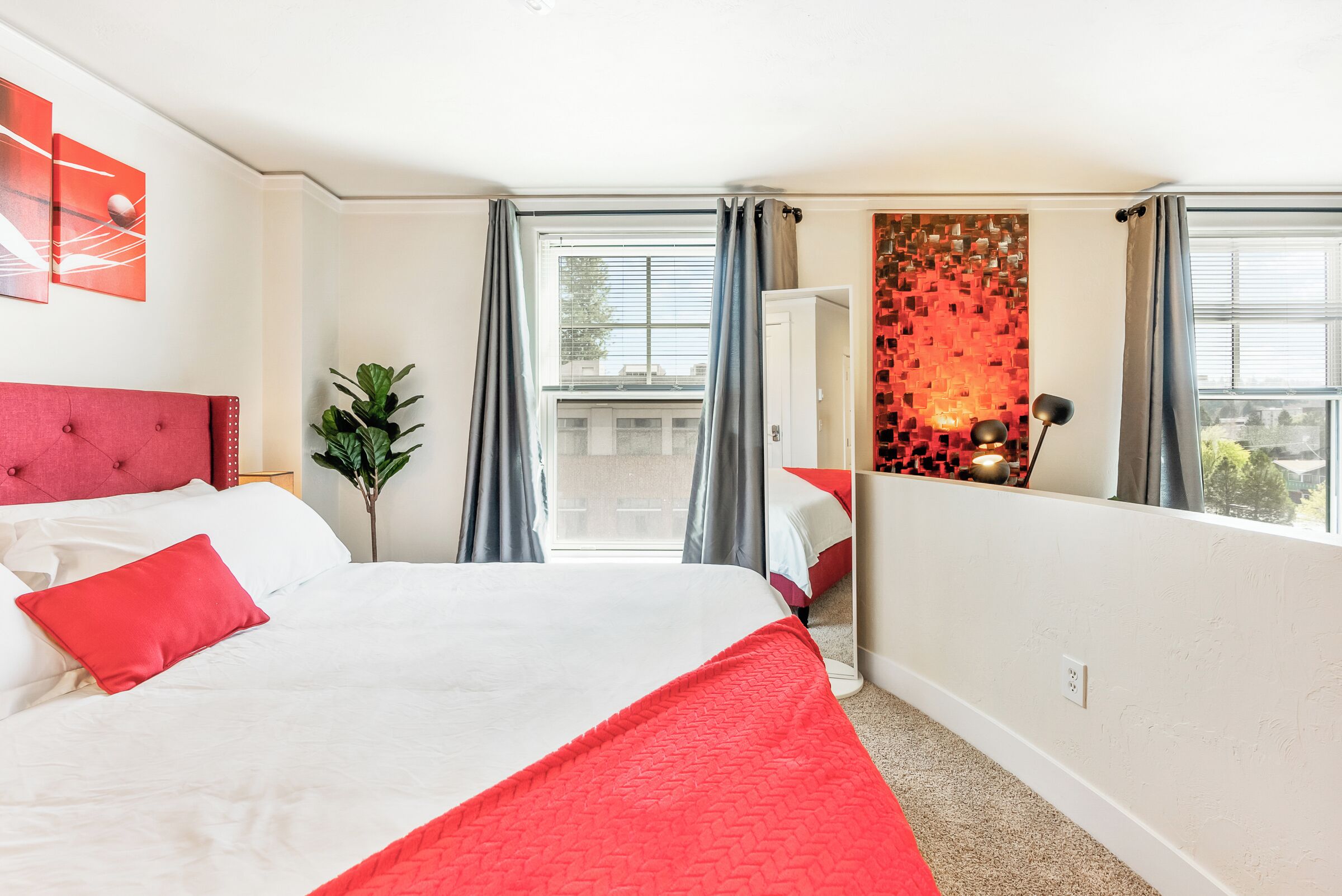 A hotel room with a large bed, a red headboard, a red throw blanket, a potted plant, a mirror, and a view of the city through the window.