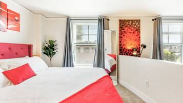 A hotel room with a large bed, a red headboard, a red throw blanket, a potted plant, a mirror, and a view of the city through the window.