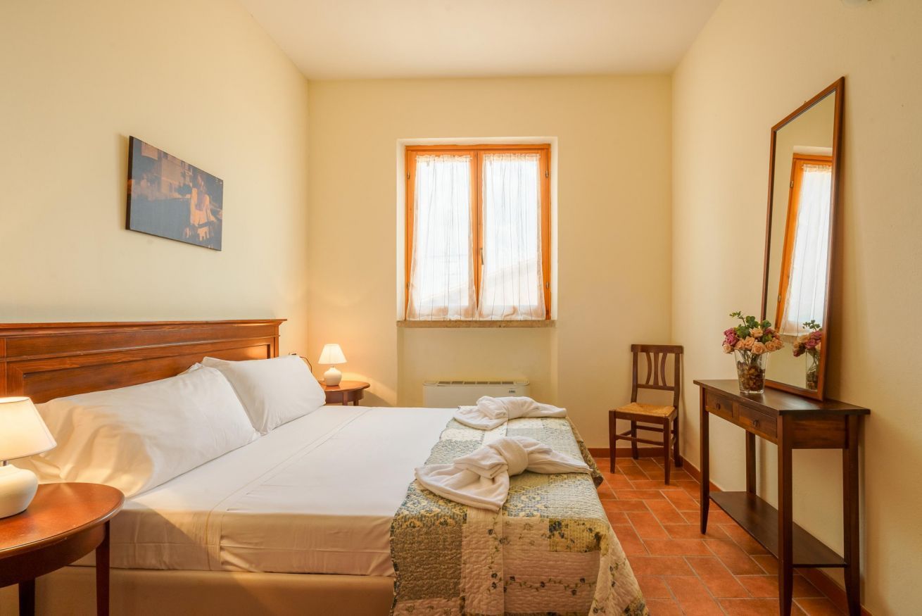 Habitación de hotel con cama, mesita de noche, una silla, escritorio, espejo y ventana con cortinas.