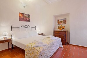 A neatly made bed with white linens and a floral bedspread, a wooden nightstand with a lamp, a dresser with a vase of flowers, and two framed pictures on the wall. - Three-room apartment on the ground or first floor in rustic style of approximately 40 m2 furnished in a simple but functional way and located in the village in a large park.It is composed of a living room with sofa and flat-screen television, kitchenette (Massa Marittima)