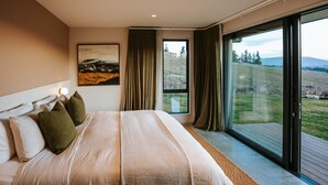 A bedroom with a large bed, a painting on the wall, and a view of a grassy landscape through a sliding glass door. - Ranger's Retreat - Secluded - Sleeps 4 (Lake Hawea)