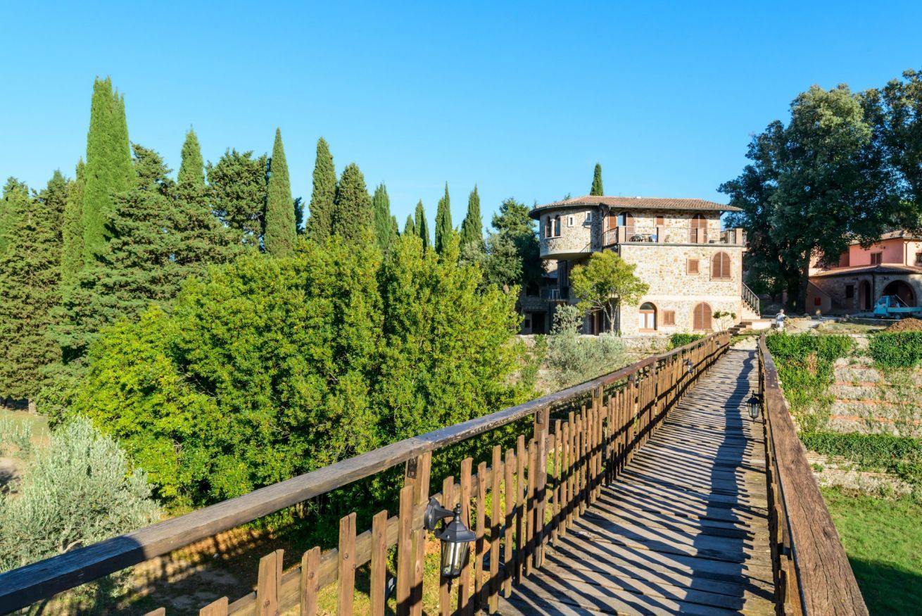 A wooden bridge leading to a stone building with a balcony, surrounded by lush greenery and tall trees.